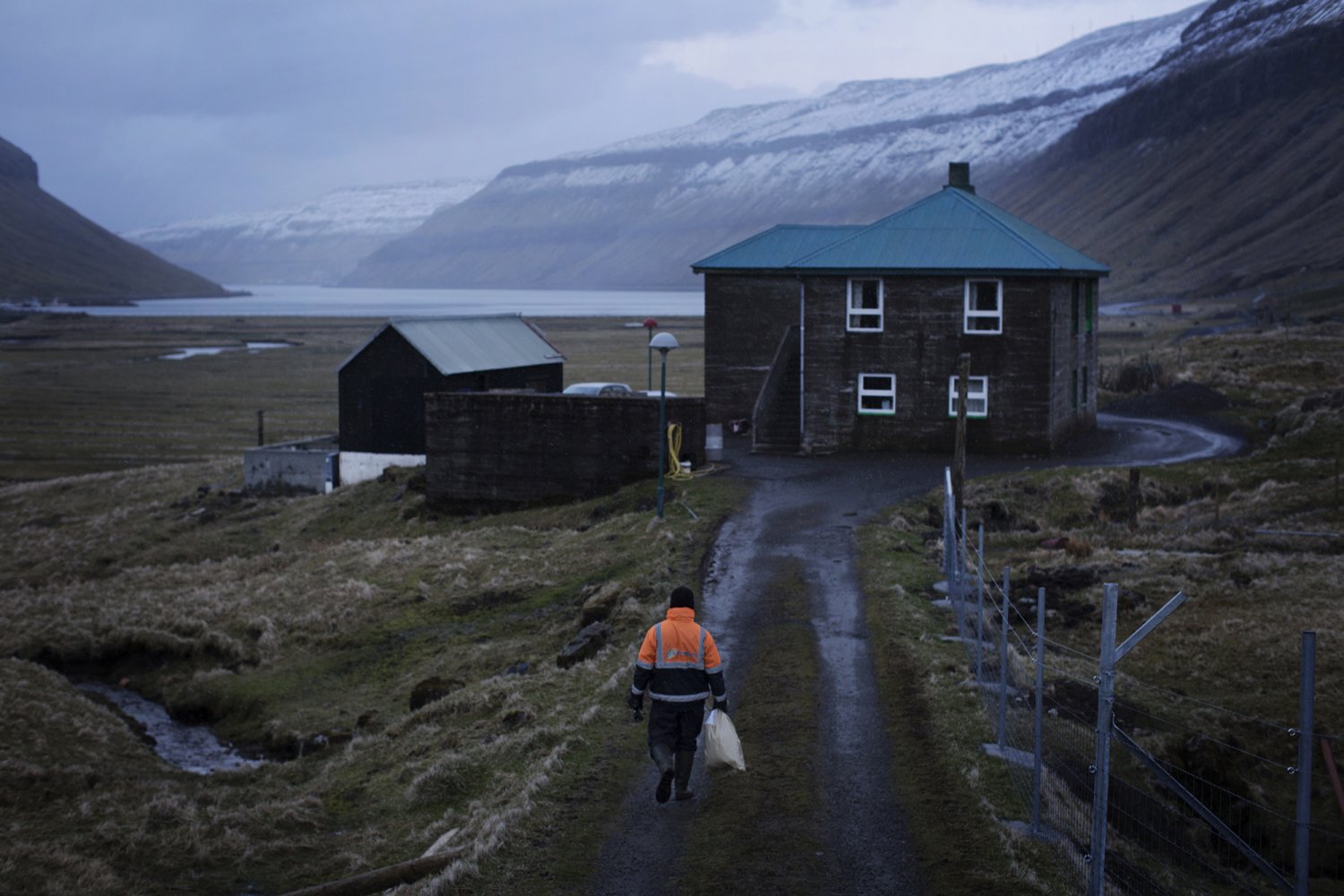 Un homme, seul et en tenue de travail, un sac à la main, avance vers une maison en bois au milieu d'un paysage magnifique, humide et froid.